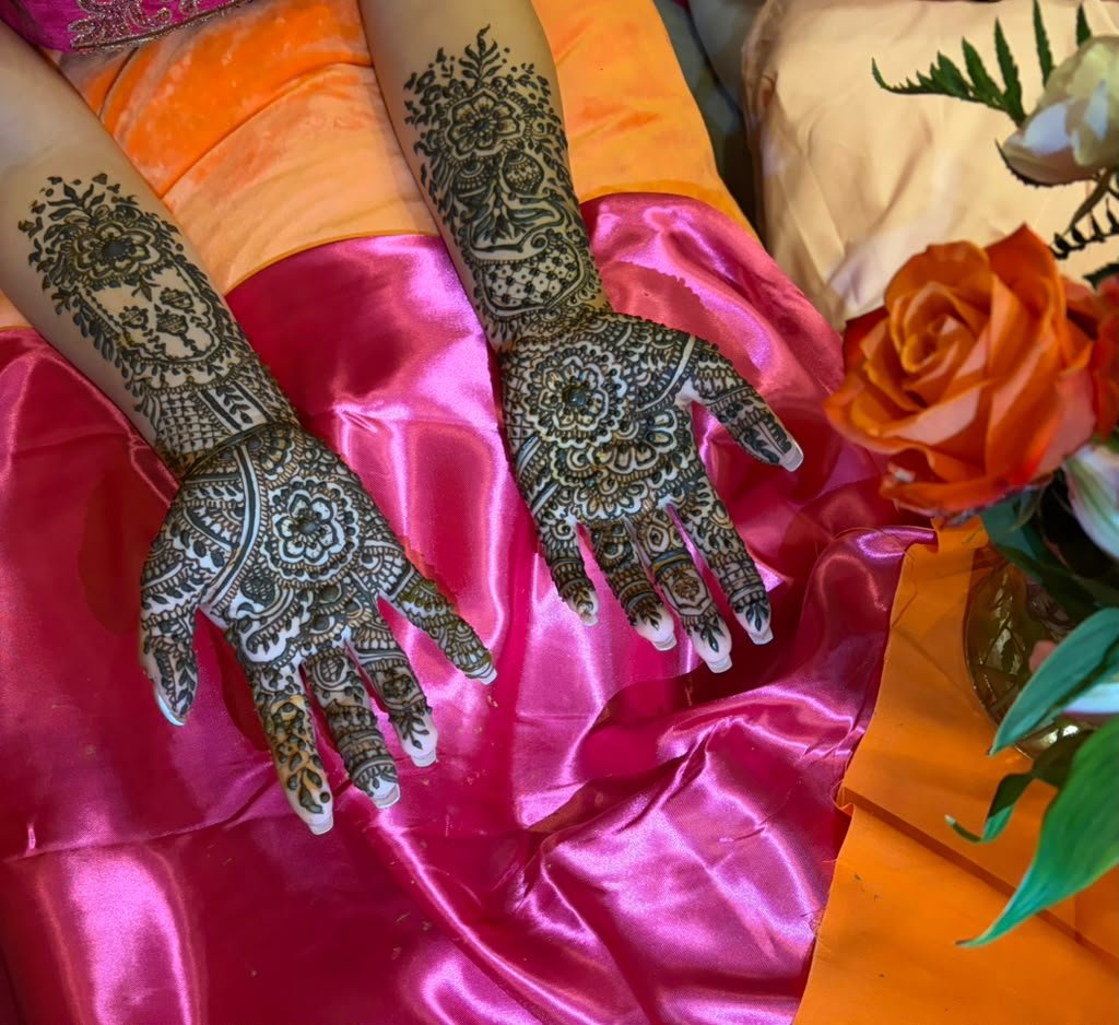 Intricate floral henna across both palms and forearms against pink and orange fabric